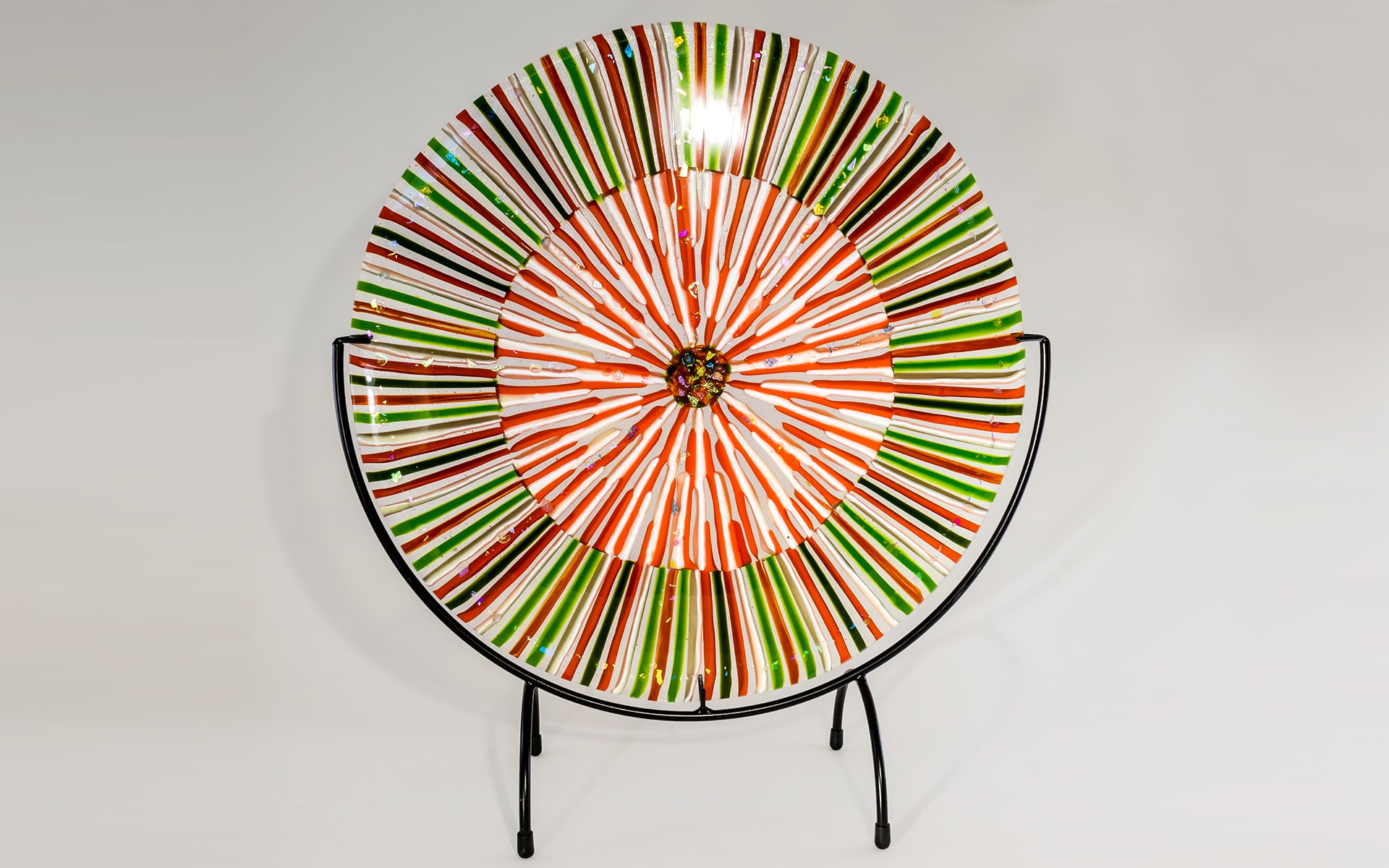 Iron Fuser 8 — large circular fused glass plate with radiating orange, green, and white stripes on a metal stand. Private collection