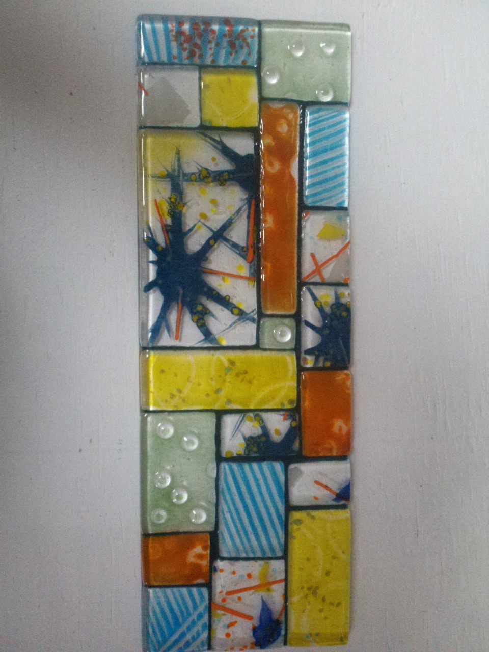 Sky Window — vertical fused glass wall panel composed of a mosaic grid of tiles in yellow, orange, teal, sage green, and white, featuring starburst patterns, stripes, and bubble accents.
